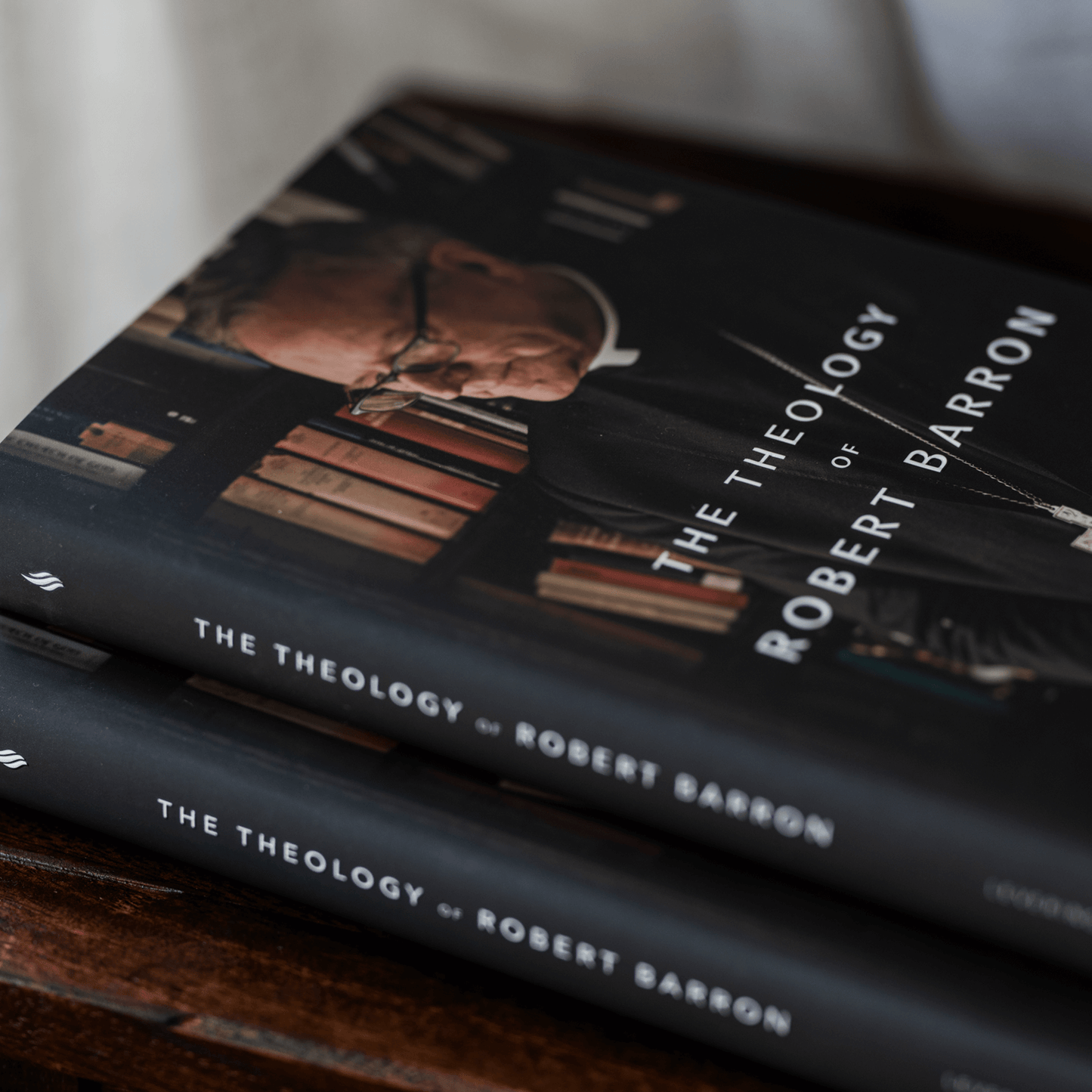 Close-up of two copies of "The Theology of Robert Barron" featuring an image of Robert Barron with glasses and collar, displayed on a wooden surface with visible bookshelves in the background, published by Word on Fire Academic, suitable for readers interested in Catholic theology books, hardcover 9781685780173
