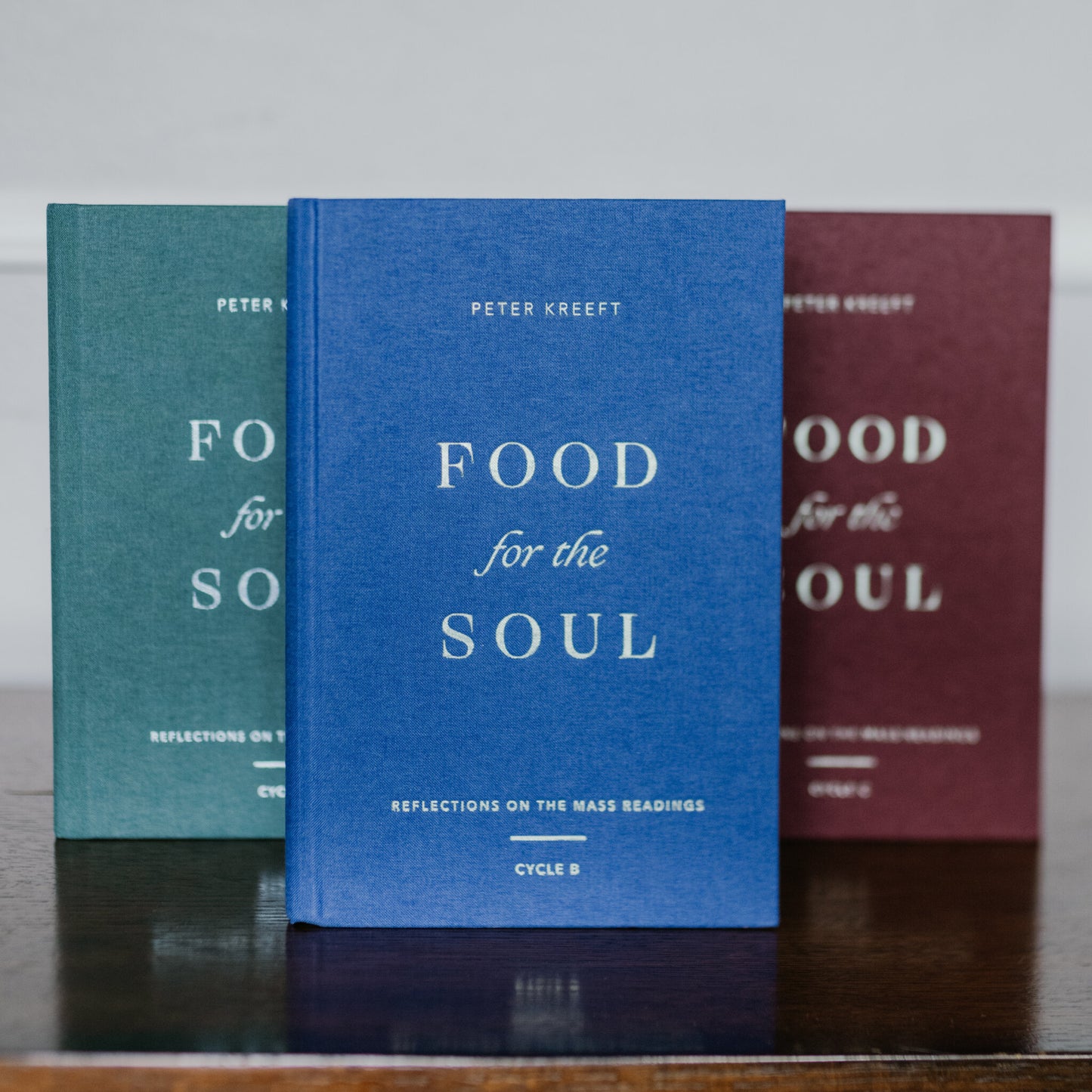 Three hardcover Catholic theology books titled "Food for the Soul - Cycle B" by Peter Kreeft in blue, green, and maroon covers displayed on a wooden surface, daily reflections on the Mass readings, suitable for prayer or Catholic gifts