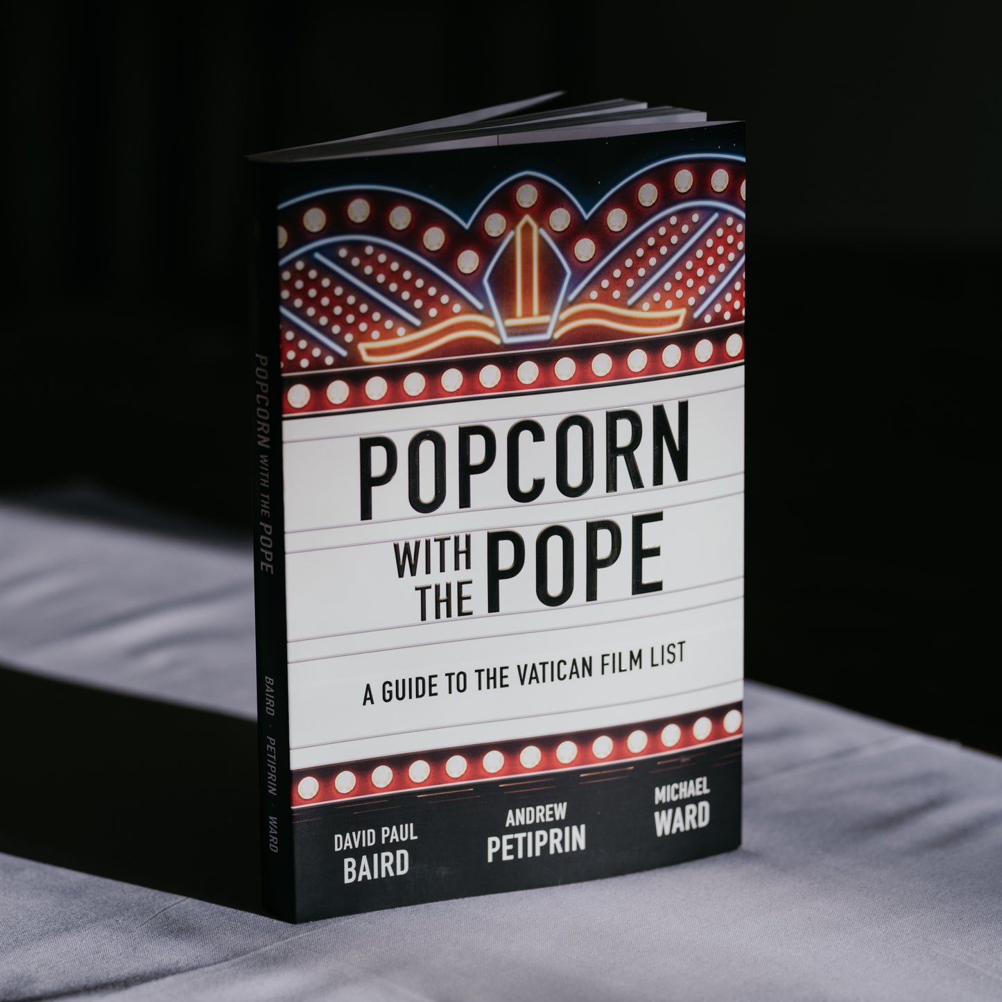 Popcorn with the Pope book standing upright with a marquee-style cover design featuring lights and a Vatican-themed graphic, by David Paul Baird, Andrew Petiprin, Michael Ward, published by Word on Fire, paperback 9781685789848
