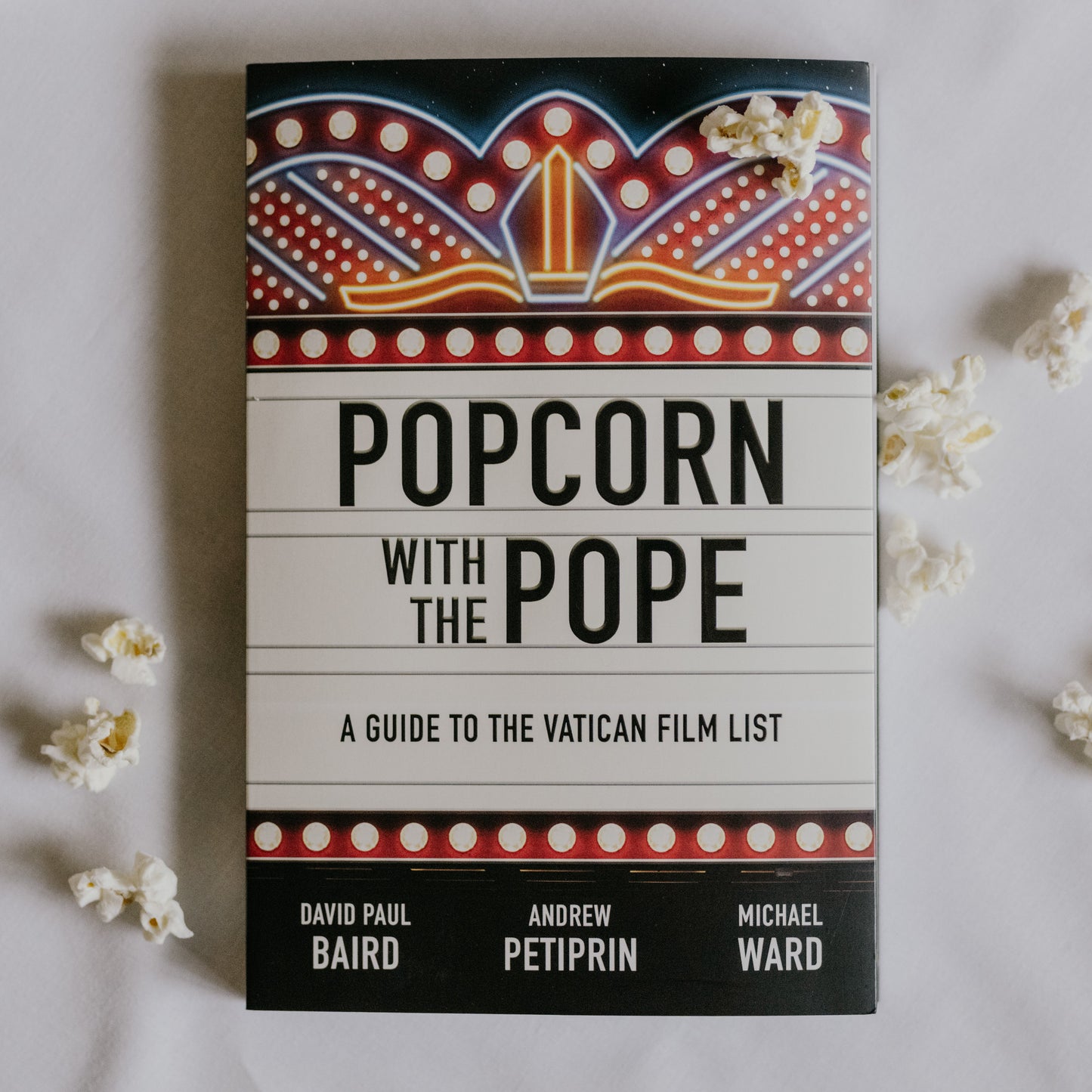 Popcorn with the Pope book cover featuring a marquee-style design with a pope's mitre symbol, surrounded by scattered popcorn, David Paul Baird, Andrew Petiprin, Michael Ward,, published by Word on Fire, paperback 9781685789848