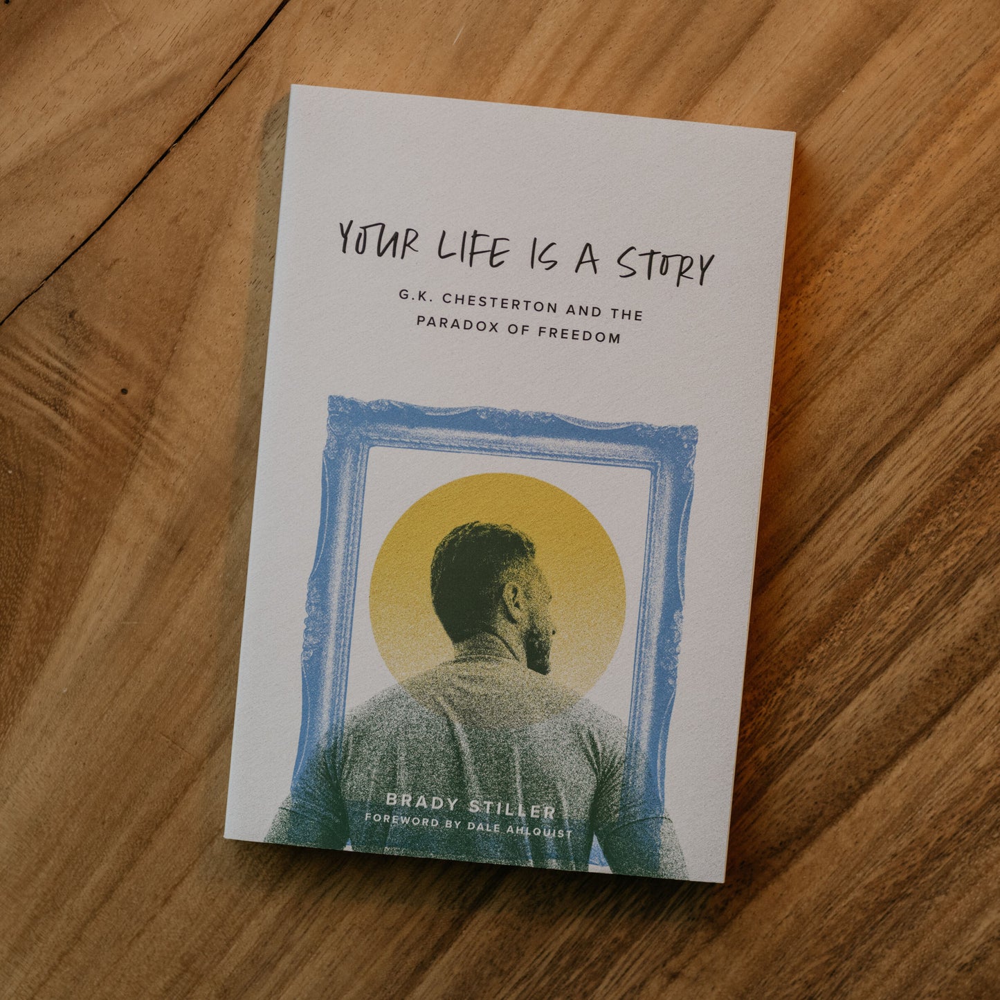 Book cover of "Your Life Is a Story" featuring a back view of a man inside a blue frame with a yellow halo-like circle behind him, with contributors G.K. Chesterton and Brady Stiller, fitting for readers interested in Catholic gifts, theology books, and spiritual growth.