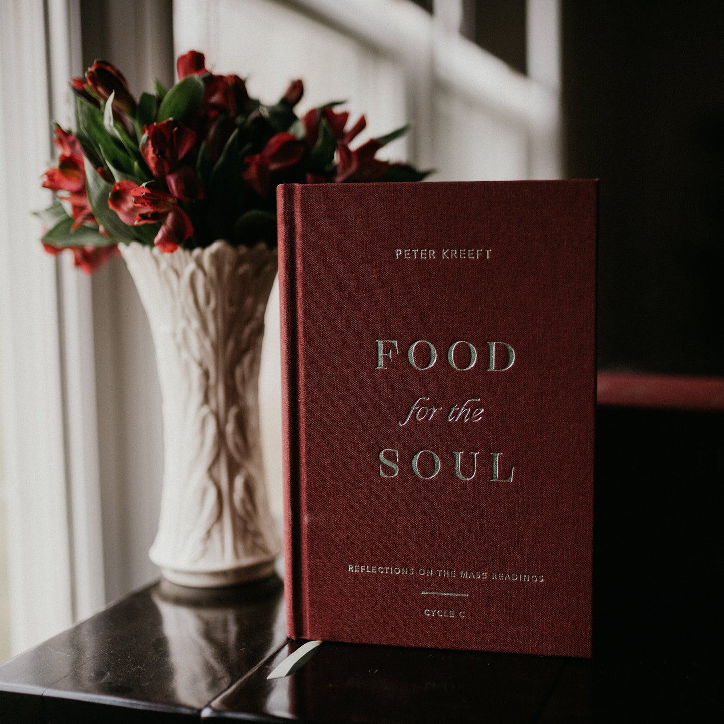 The hardcover book "Food for the Soul - Cycle C" by Peter Kreeft displayed next to a vase of red flowers, book for spiritual growth and Catholic reflection, by Peter Kreeft, reflections on the daily Mass readings