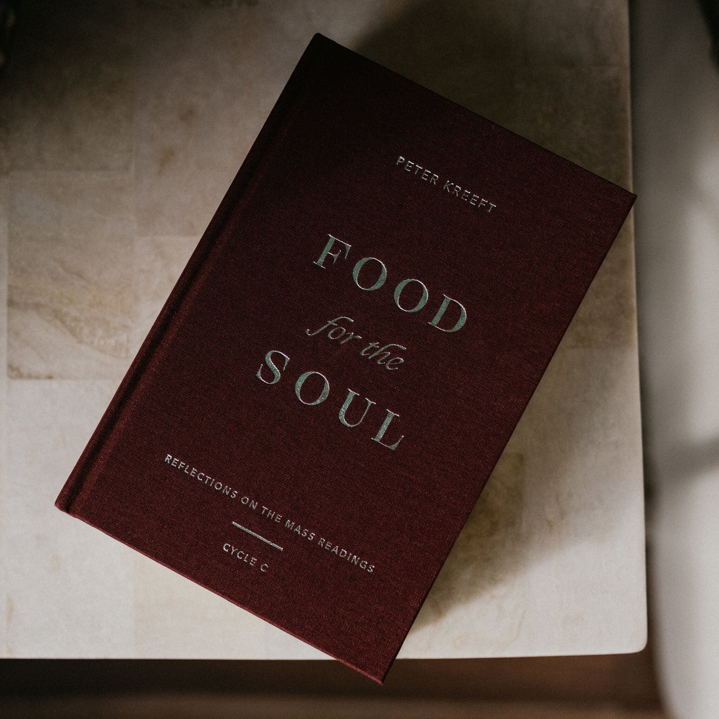 A maroon hardcover book titled "Food for the Soul - Cycle C" by Peter Kreeft rests on a light-colored surface, featuring reflections on the daily Mass readings for spiritual growth and evangelization