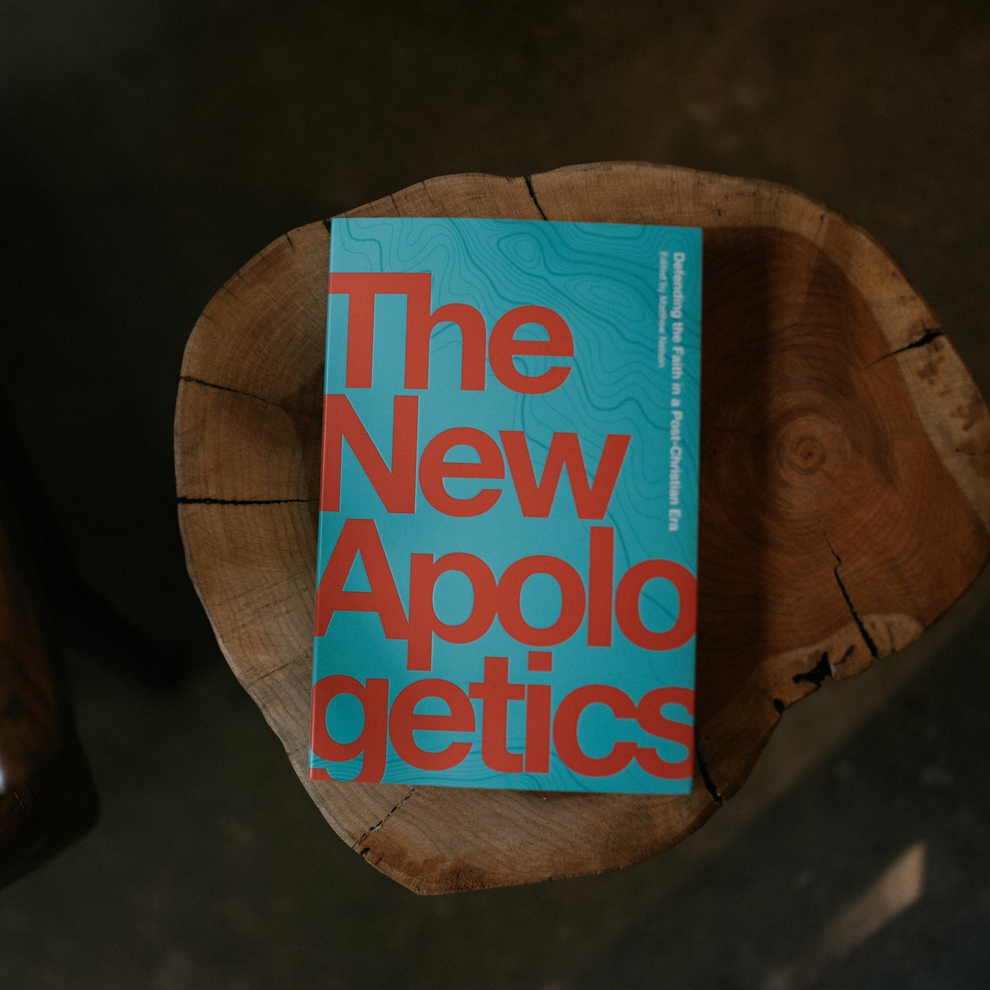 The New Apologetics book with bold red text on a blue cover placed on a rustic wooden surface, suitable for spiritual growth and evangelization, authored by multiple contributors including Matthew Nelson and Thomas Collins, paperback 9781685780043