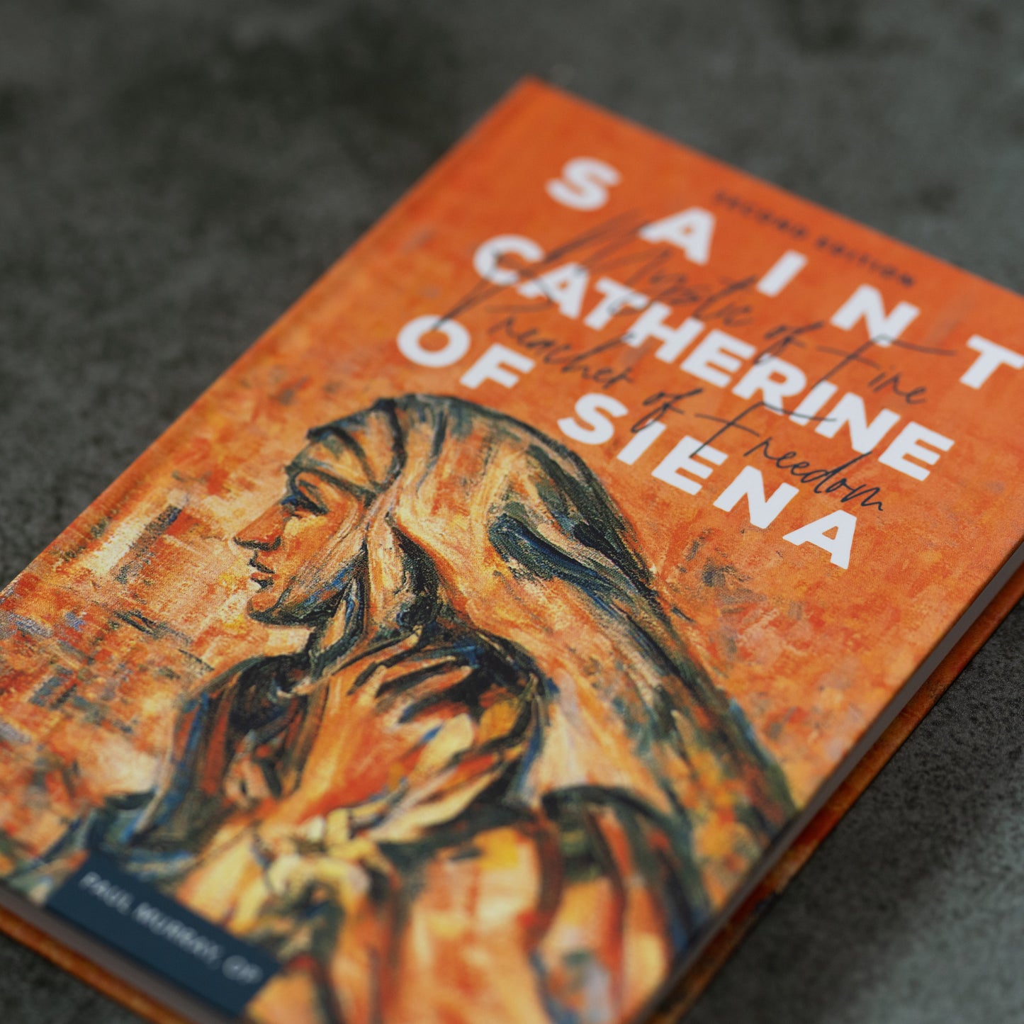 An artistic close-up of the book Saint Catherine of Siena with an orange cover featuring a painted portrait of the saint, by Father Paul Murray, OP, reflections on St. Catherine of Siena, book on Catholic female saint, hardcover 9781943243648