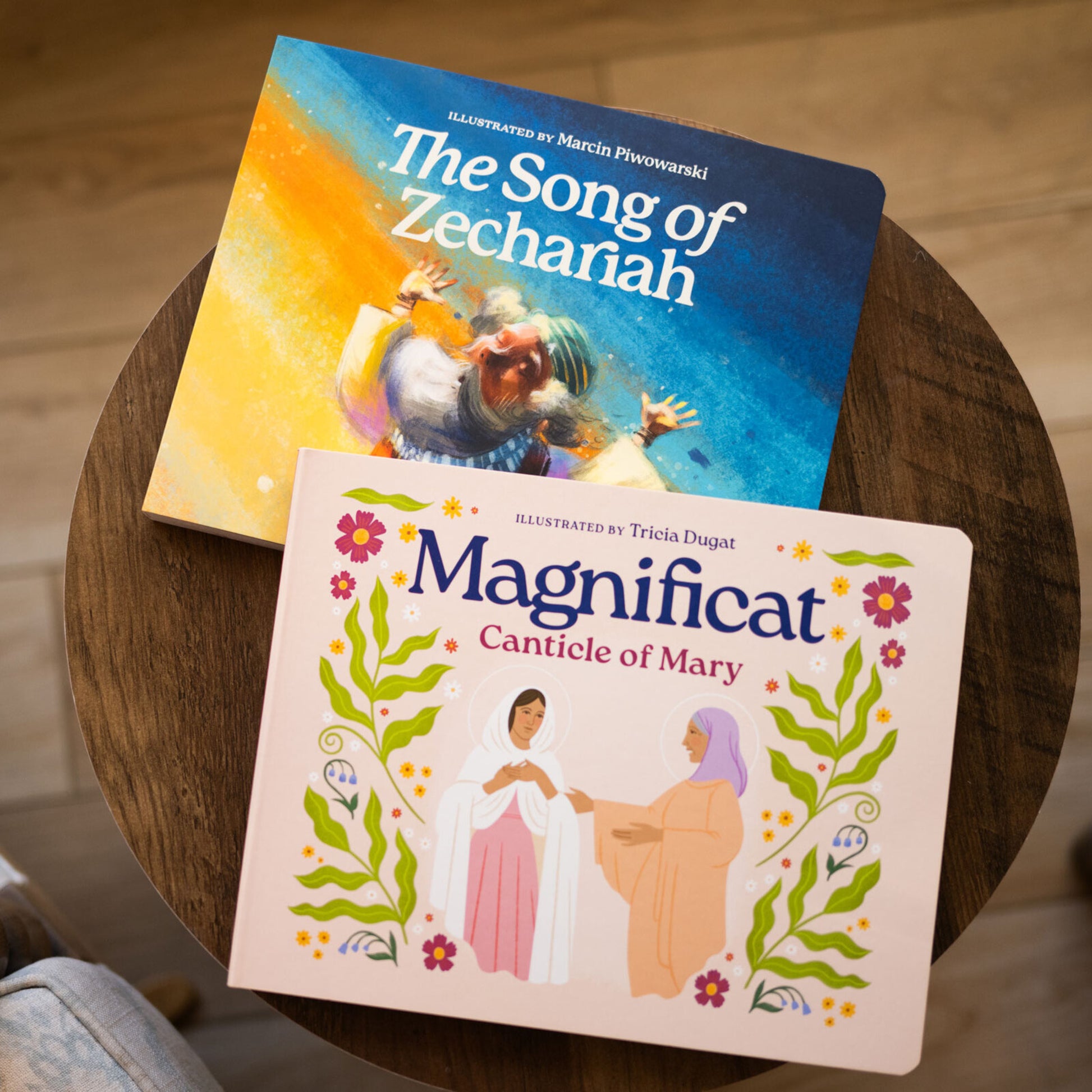 Two illustrated Catholic children’s books lying on a wooden table, titled The Song of Zechariah and Magnificat Canticle of Mary, featuring vibrant cover art, for ages 1-5, helping babies and toddlers pray with scripture